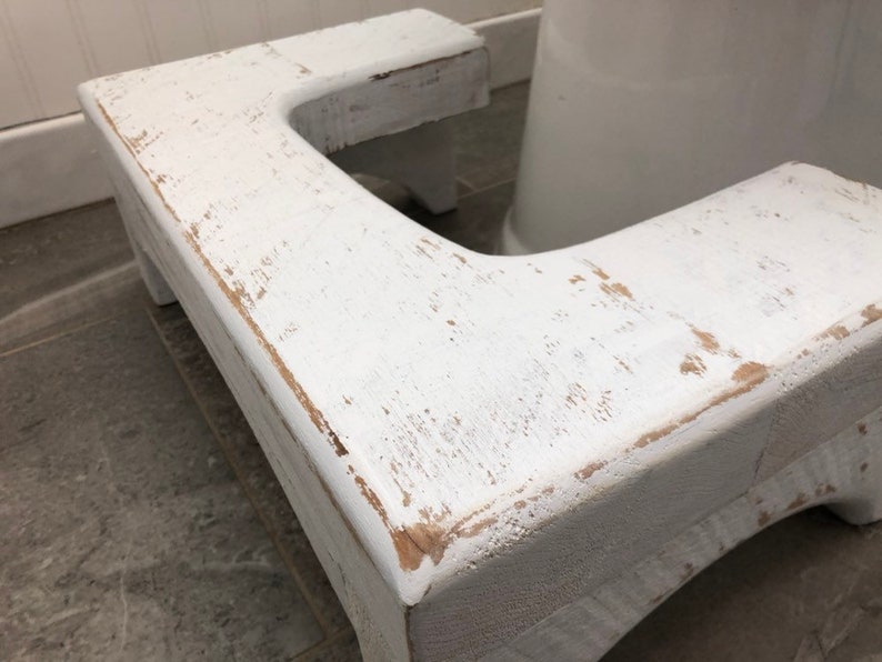 May include: White wooden toilet seat riser with a distressed finish. The riser is designed to help people reach the toilet seat more easily.