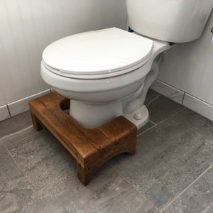 May include: A wooden step stool with a rounded cutout for a toilet base. The stool is made of dark brown wood and has a natural finish. The stool is placed in front of a white toilet.