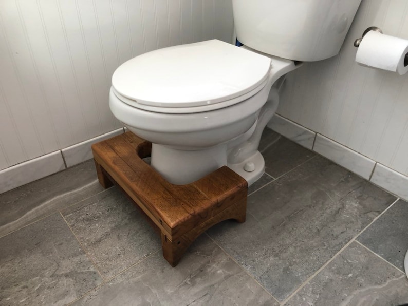 May include: A wooden step stool with a rounded cutout for a toilet base. The stool is made of dark brown wood and has a natural finish. The stool is placed in front of a white toilet.