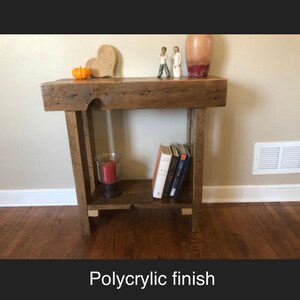 May include: A rustic wooden side table with a shelf, featuring a polycrylic finish. The table has a heart-shaped decorative object, a small orange pumpkin, and two figurines on top. There are books and a candle on the shelf.