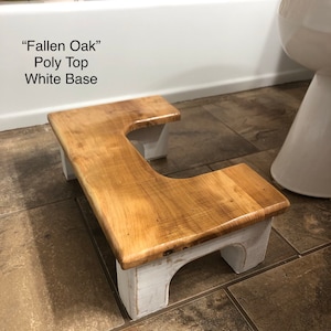 May include: A wooden bathroom step stool with a white base and a natural wood top. The top is shaped like a U and has a "Fallen Oak" Poly Top White Base label.