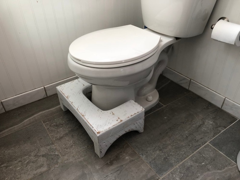 May include: White toilet with a white wooden step stool in a bathroom with gray tile flooring.