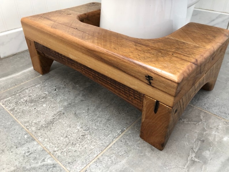 May include: A wooden toilet seat riser with a curved front edge and a natural wood finish. The riser is made of reclaimed wood and has a rustic look.