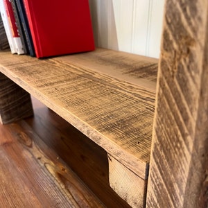 May include: A rustic wooden shelf with a red book and other books on the top shelf. The shelf is made of reclaimed wood and has a natural, weathered finish.