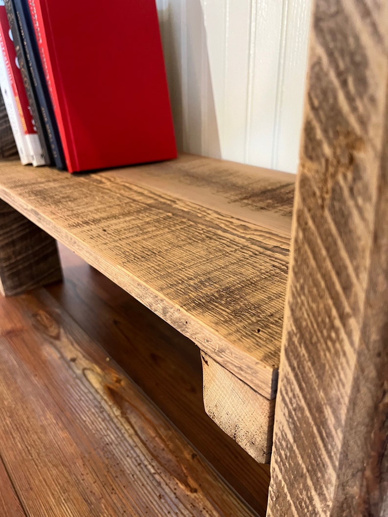 May include: A rustic wooden shelf with a red book and other books on the top shelf. The shelf is made of reclaimed wood and has a natural, weathered finish.