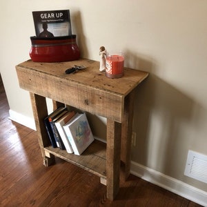 May include: A rustic wooden console table with a shelf, featuring a red ceramic bowl with a book titled "Gear Up" inside, a small decorative figurine, and a candle. The table is made of reclaimed wood and has a natural finish.
