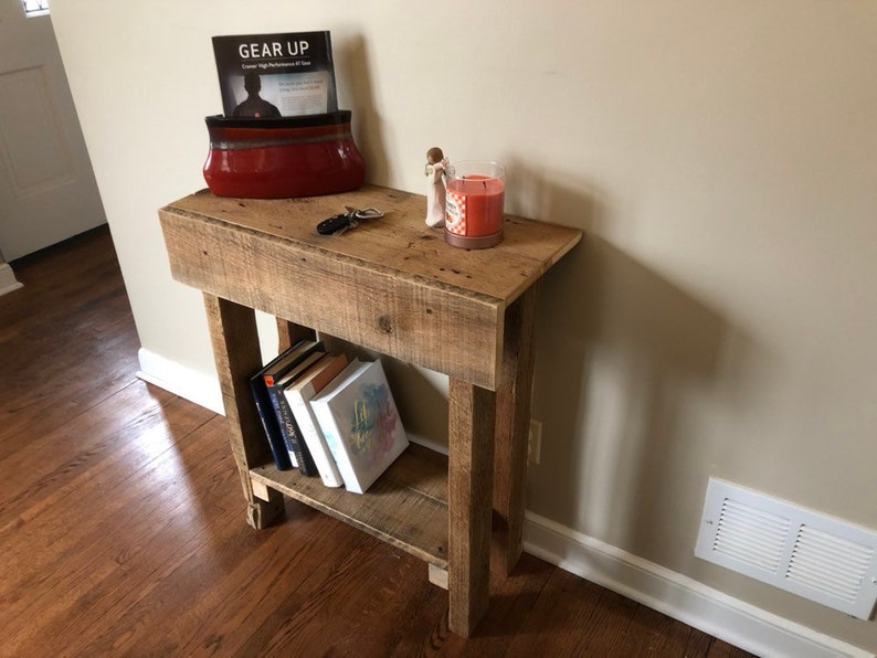 May include: A rustic wooden console table with a shelf, featuring a red ceramic bowl with a book titled "Gear Up" inside, a small decorative figurine, and a candle. The table is made of reclaimed wood and has a natural finish.