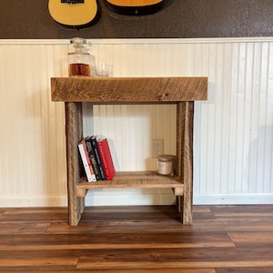 May include: A rustic wooden console table with a shelf, featuring a distressed brown finish. The table has a single shelf and is decorated with books and a candle. Two acoustic guitars hang on the wall behind the table.