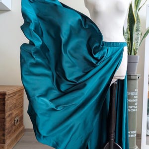 May include: A floor-length teal skirt made of a smooth, satin-like fabric. The skirt features a high-waisted design with an elastic waistband and a dramatic, flowing silhouette that flares out to one side, showcasing the fabric's sheen.