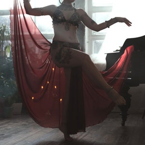 May include: A dark-colored, embellished bra top with intricate metal detailing and a matching hip belt, paired with a long, flowing, sheer deep red skirt that drapes from the waist, featuring metallic arm cuffs and a decorative neck piece.