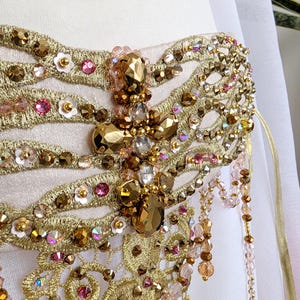 May include: A pale pink bodice featuring intricate gold metallic embroidery, embellished with faceted amber-toned crystals, small pink sequins, and white floral accents. Cascading beaded fringe in shades of pink and gold hangs from the side.