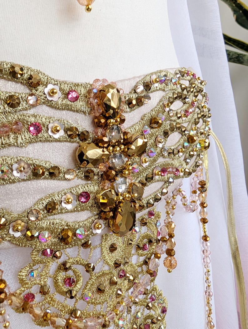 May include: A pale pink bodice featuring intricate gold metallic embroidery, embellished with faceted amber-toned crystals, small pink sequins, and white floral accents. Cascading beaded fringe in shades of pink and gold hangs from the side.