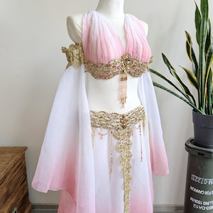 May include: A two-piece dance costume featuring a white and pink ombre chiffon top and skirt. The top has a V-neckline and off-the-shoulder sleeves, adorned with gold lace embroidery, sequins, and dangling beaded fringe accents at the center.