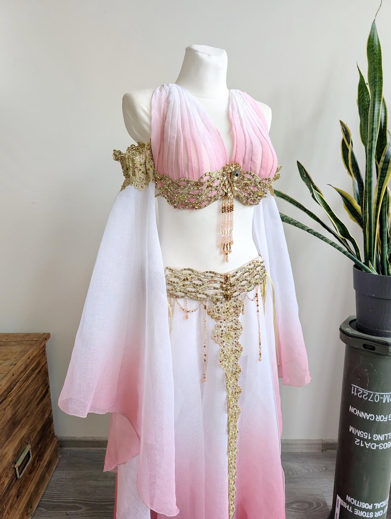 May include: A two-piece dance costume featuring a white and pink ombre chiffon top and skirt. The top has a V-neckline and off-the-shoulder sleeves, adorned with gold lace embroidery, sequins, and dangling beaded fringe accents at the center.