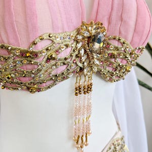 May include: A light pink pleated top featuring an intricate gold embroidered overlay adorned with clear crystals, metallic sequins, and small pink beads. A central cluster of larger crystals hangs with delicate strands of light pink and gold beads.