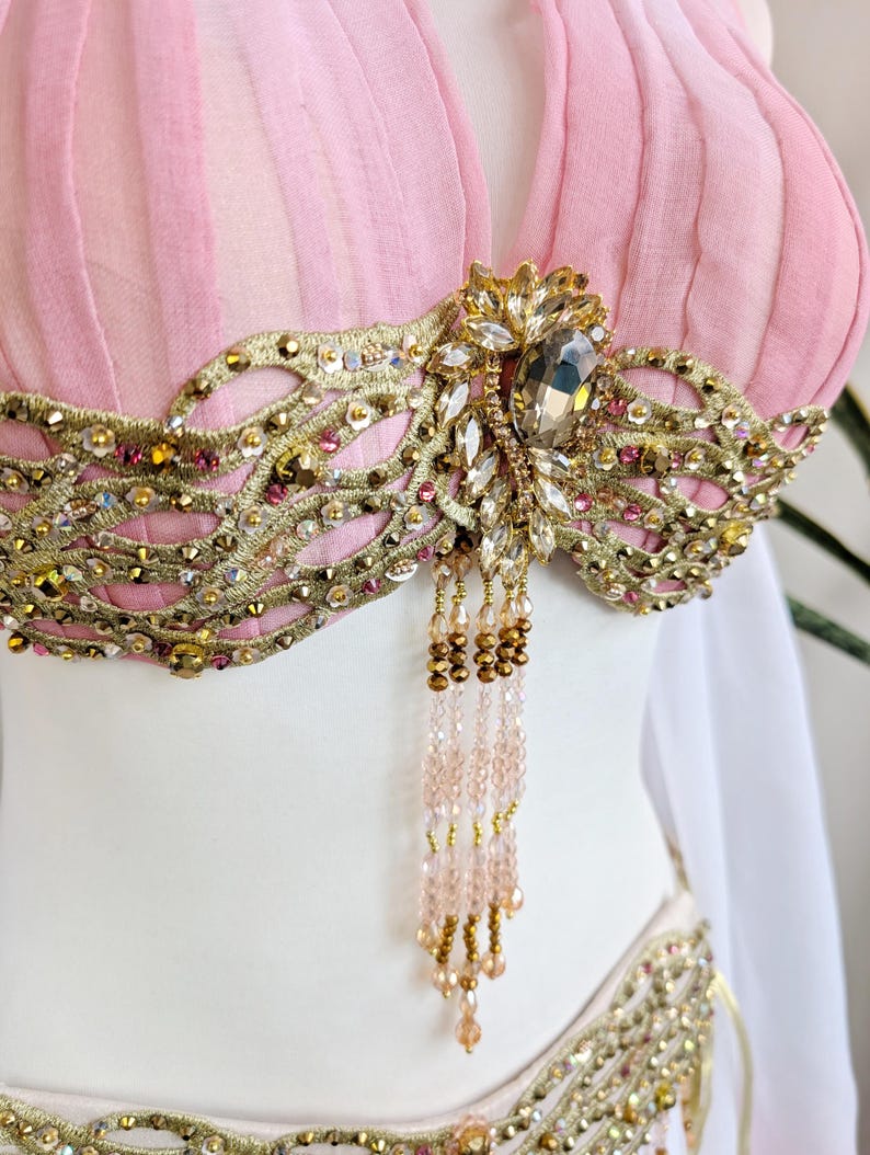 May include: A light pink pleated top featuring an intricate gold embroidered overlay adorned with clear crystals, metallic sequins, and small pink beads. A central cluster of larger crystals hangs with delicate strands of light pink and gold beads.