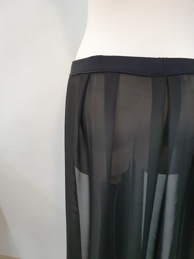 May include: A black skirt featuring a sheer, semi-transparent fabric with vertical pleated panels. The garment has a solid, opaque waistband and a flowing, lightweight construction that hangs straight down from the waistline.