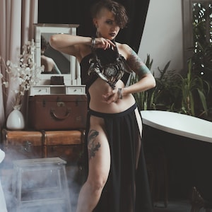 May include: A black halter top with a cutout midriff and a matching black floor-length skirt featuring high side slits. The outfit is paired with a large, dark, intricate statement necklace featuring horn-like elements and a central circular pendant.