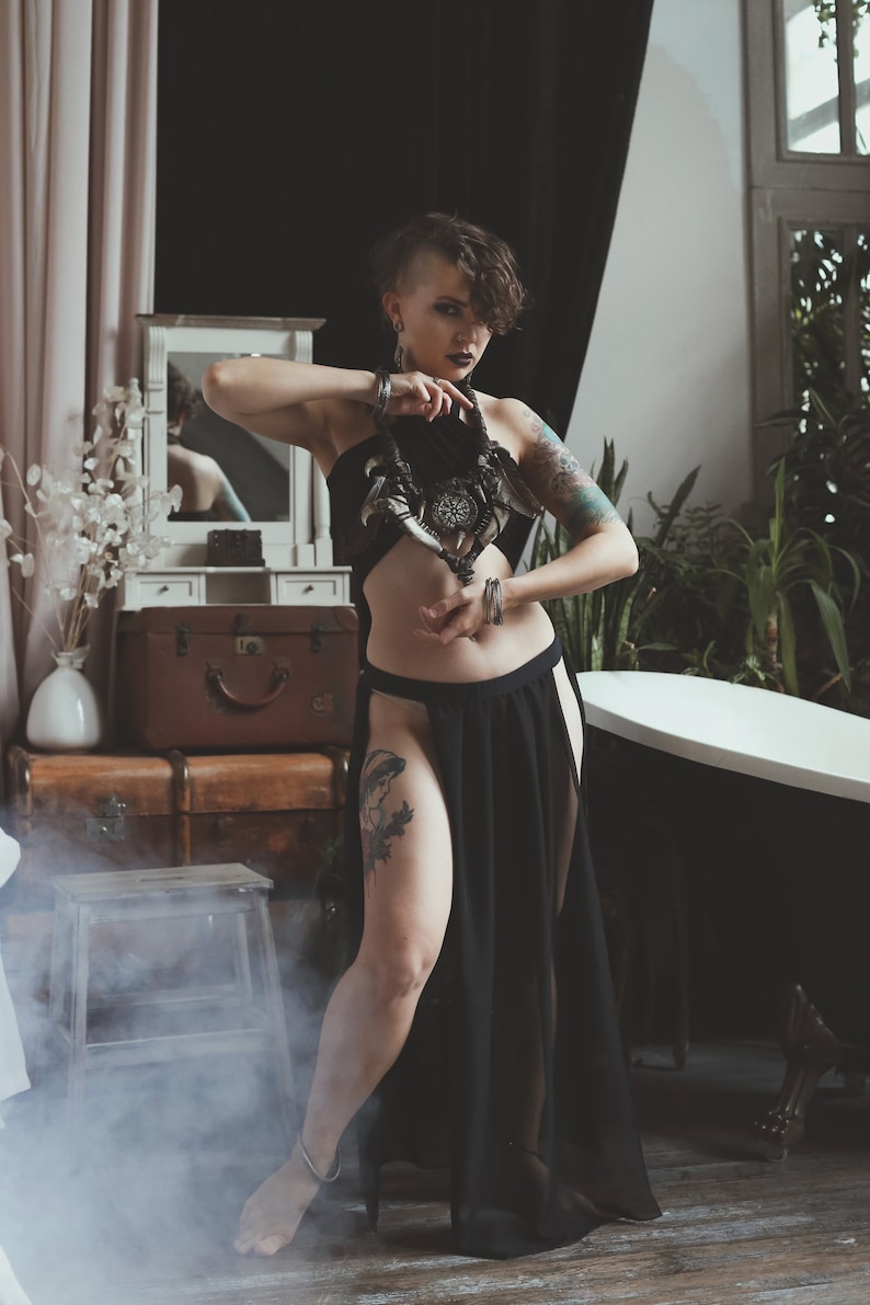 May include: A black halter top with a cutout midriff and a matching black floor-length skirt featuring high side slits. The outfit is paired with a large, dark, intricate statement necklace featuring horn-like elements and a central circular pendant.