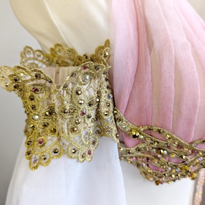 May include: A garment featuring a pale pink pleated bodice and a white sleeve, accented with intricate gold lace trim. The lace is embellished with small, reflective crystals in shades of gold and pink, creating a textured, ornate appearance.