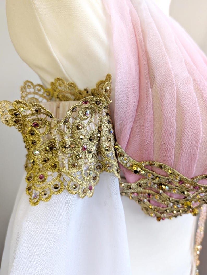 May include: A garment featuring a pale pink pleated bodice and a white sleeve, accented with intricate gold lace trim. The lace is embellished with small, reflective crystals in shades of gold and pink, creating a textured, ornate appearance.