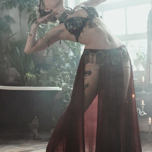 May include: A dark burgundy sheer skirt with a high-cut design, paired with a black embellished bra top featuring metallic accents and dangling charms. The outfit includes a decorative waist belt with circular metal details and hanging fringe elements.