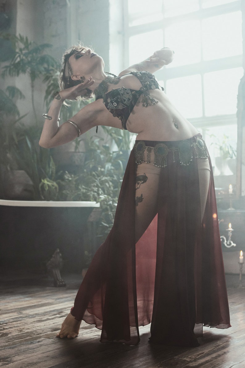 May include: A dark burgundy sheer skirt with a high-cut design, paired with a black embellished bra top featuring metallic accents and dangling charms. The outfit includes a decorative waist belt with circular metal details and hanging fringe elements.