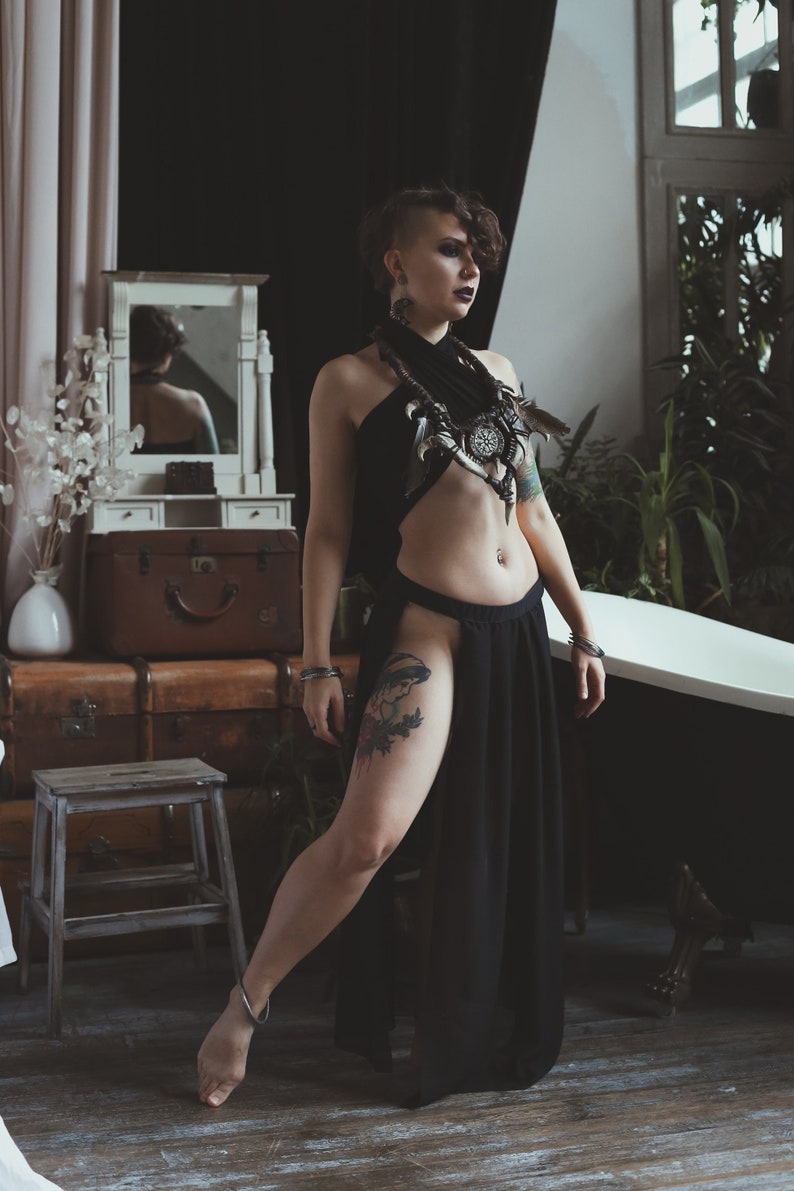 May include: A black halter-style top paired with a matching high-slit skirt. The outfit features a decorative neckpiece adorned with feathers, dark metallic accents, and a central circular emblem. The skirt has a high waistband and long, flowing fabric.