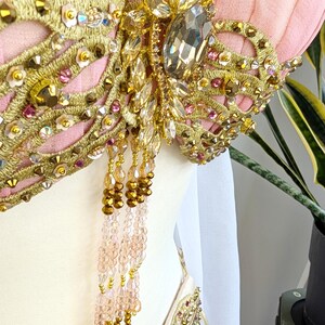 May include: A light pink garment featuring intricate gold embroidery, clear and pink faceted crystals, and metallic sequins. A central cluster of large clear stones is accented by dangling strands of gold and pale pink beads hanging from the bodice.