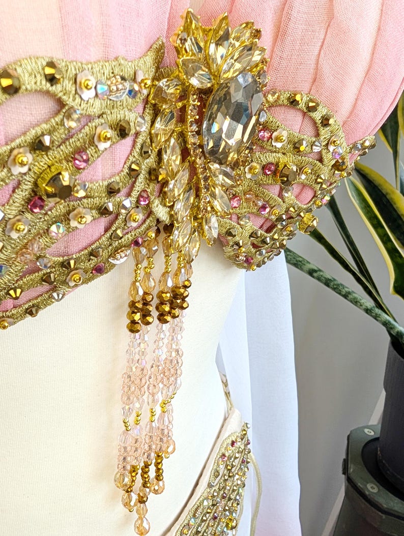 May include: A light pink garment featuring intricate gold embroidery, clear and pink faceted crystals, and metallic sequins. A central cluster of large clear stones is accented by dangling strands of gold and pale pink beads hanging from the bodice.