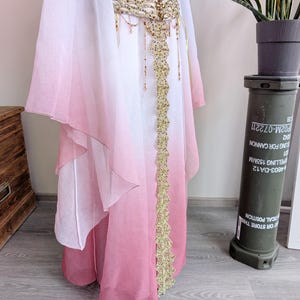 May include: A floor-length skirt featuring a white-to-pink ombre gradient, paired with a matching sheer cape. The skirt is accented by a wide, gold-toned belt with intricate beadwork and a vertical gold lace panel extending down the front center.