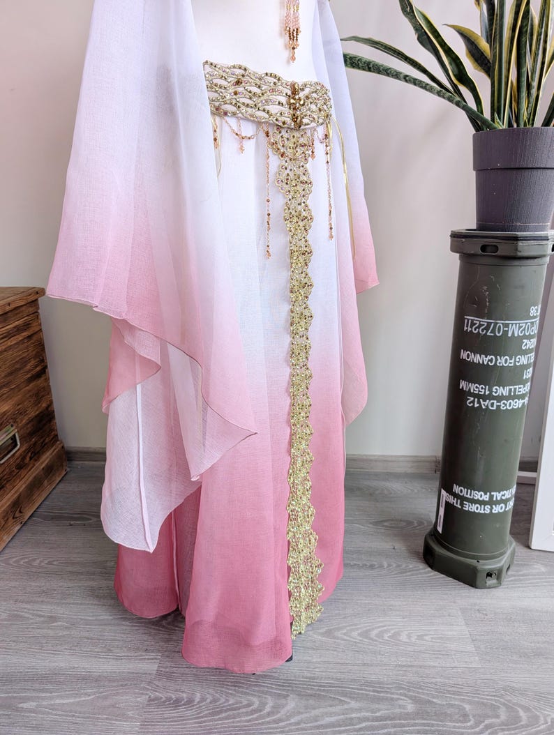 May include: A floor-length skirt featuring a white-to-pink ombre gradient, paired with a matching sheer cape. The skirt is accented by a wide, gold-toned belt with intricate beadwork and a vertical gold lace panel extending down the front center.