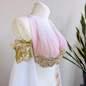 May include: A sleeveless bodice featuring pleated light pink fabric, adorned with intricate gold lace trim, metallic embroidery, and small clear crystals. The garment includes detached off-the-shoulder sleeves made of sheer white fabric.