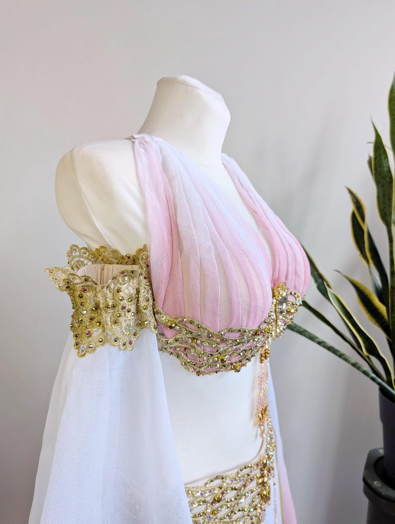 May include: A sleeveless bodice featuring pleated light pink fabric, adorned with intricate gold lace trim, metallic embroidery, and small clear crystals. The garment includes detached off-the-shoulder sleeves made of sheer white fabric.