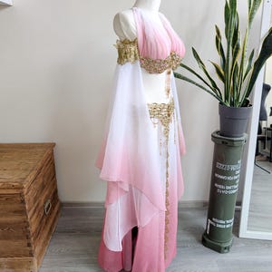 May include: A two-piece costume featuring a pink and white ombre chiffon top and skirt. The top has off-the-shoulder sleeves and gold beaded embellishments. The skirt includes a matching gold beaded waistband with hanging fringe details.
