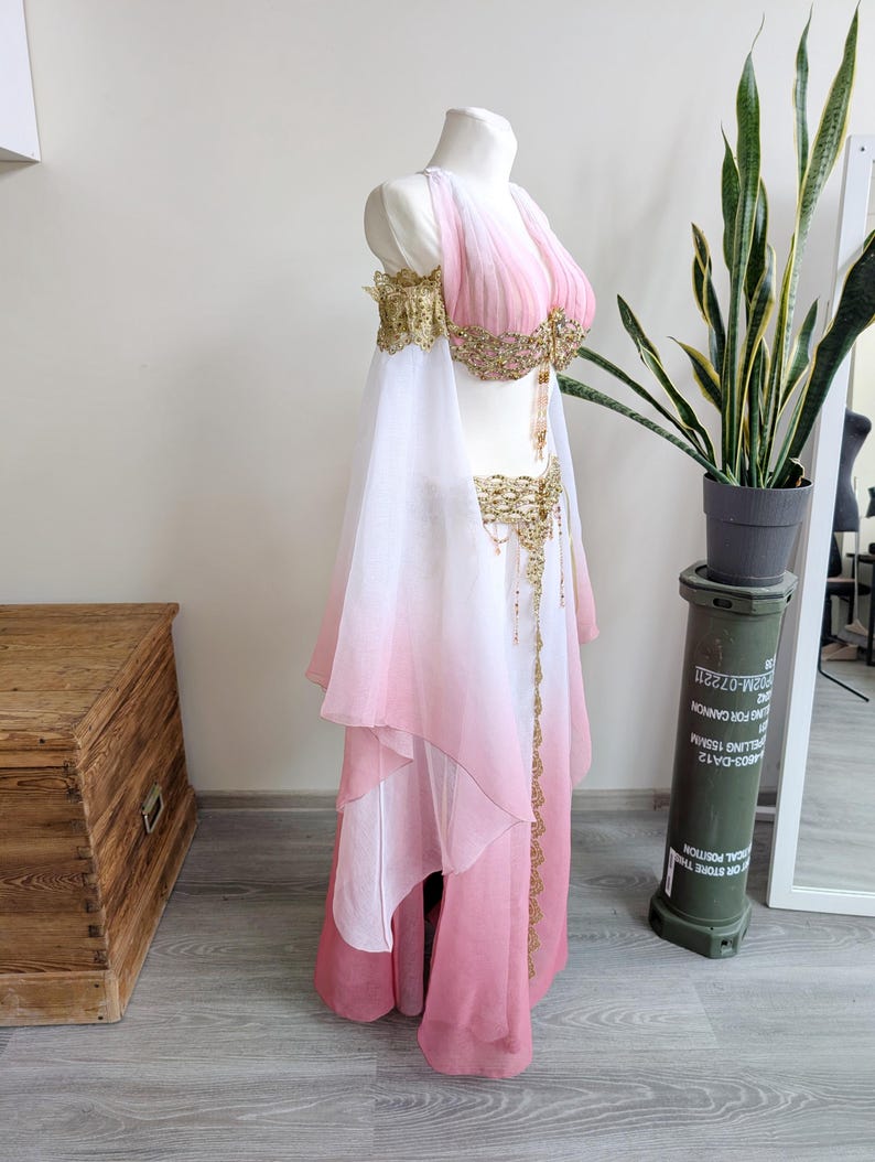 May include: A two-piece costume featuring a pink and white ombre chiffon top and skirt. The top has off-the-shoulder sleeves and gold beaded embellishments. The skirt includes a matching gold beaded waistband with hanging fringe details.
