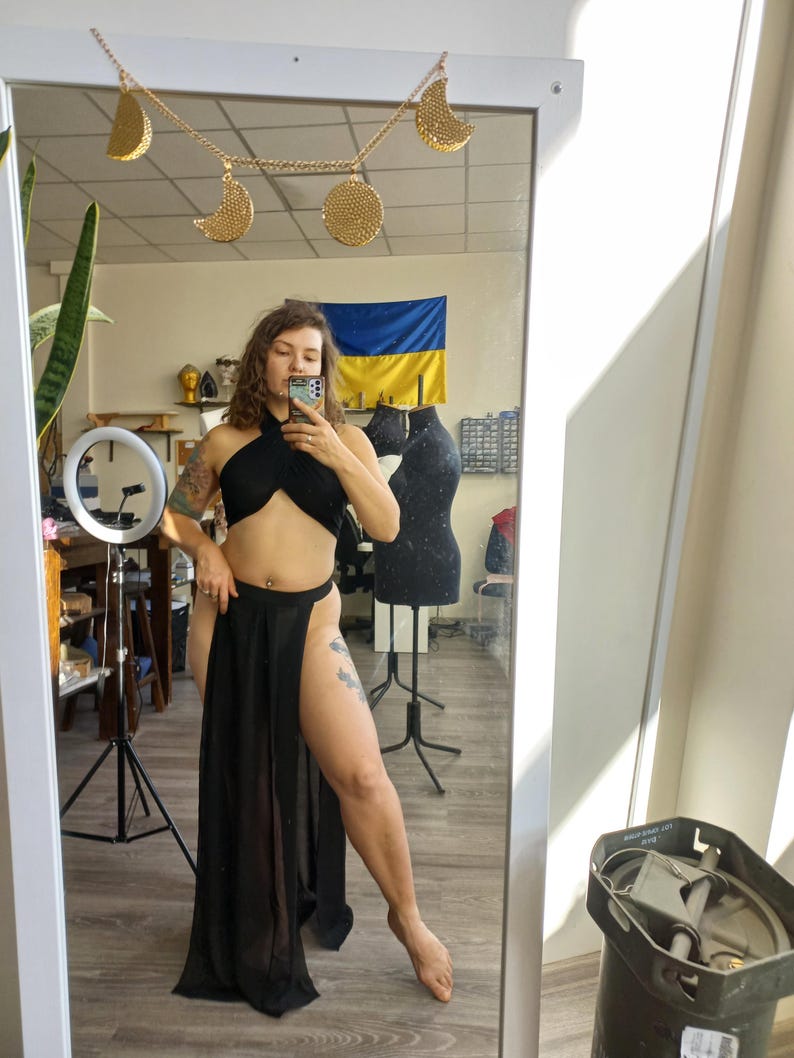 May include: A black two-piece outfit consisting of a halter-style crop top with a crisscross front and a high-waisted, floor-length skirt featuring a sheer, flowing fabric with a high side slit, reflected in a mirror with a gold moon phase garland.