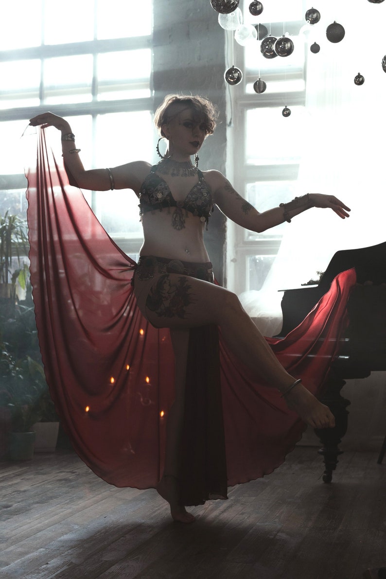 May include: A dark-colored, embellished bra top with intricate metal detailing and a matching hip belt, paired with a long, flowing, sheer deep red skirt that drapes from the waist, featuring metallic arm cuffs and a decorative neck piece.