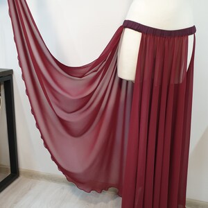 May include: A floor-length, sheer burgundy skirt featuring a dark maroon elastic waistband. The skirt is constructed from a lightweight, translucent fabric with a flowing, draped design that extends to one side, showcasing a scalloped hemline.