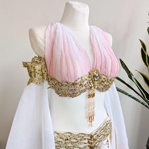 May include: A two-piece costume featuring a pink and white pleated bodice with gold metallic embroidery, intricate beadwork, and dangling crystal accents. The set includes matching gold-trimmed arm bands and a flowing white skirt with gold detailing.