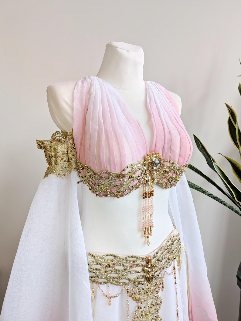 May include: A two-piece costume featuring a pink and white pleated bodice with gold metallic embroidery, intricate beadwork, and dangling crystal accents. The set includes matching gold-trimmed arm bands and a flowing white skirt with gold detailing.