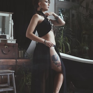 May include: A black two-piece outfit consisting of a sleeveless halter crop top and a long, flowing skirt with a high side slit. The skirt features a sheer overlay, and the ensemble is paired with silver-toned jewellery including a necklace and bracelets.