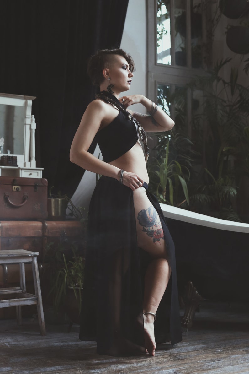 May include: A black two-piece outfit consisting of a sleeveless halter crop top and a long, flowing skirt with a high side slit. The skirt features a sheer overlay, and the ensemble is paired with silver-toned jewellery including a necklace and bracelets.