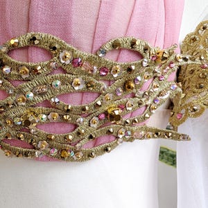 May include: A light pink garment featuring a wide, horizontal band of intricate gold metallic embroidery. The gold trim is embellished with numerous small, sparkling clear and pink crystals, along with tiny white floral accents throughout.
