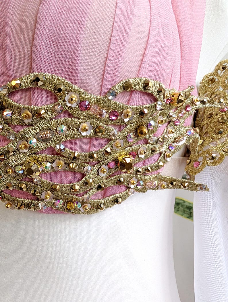 May include: A light pink garment featuring a wide, horizontal band of intricate gold metallic embroidery. The gold trim is embellished with numerous small, sparkling clear and pink crystals, along with tiny white floral accents throughout.