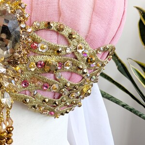 May include: A pink fabric bodice featuring intricate gold embroidery in a swirling pattern. The design is embellished with numerous small gold studs, iridescent sequins, tiny white flower appliqu&eacute;s, and pink crystals. A large faceted amber-colored crystal sits at the center, with dangling beaded strands hanging below.
