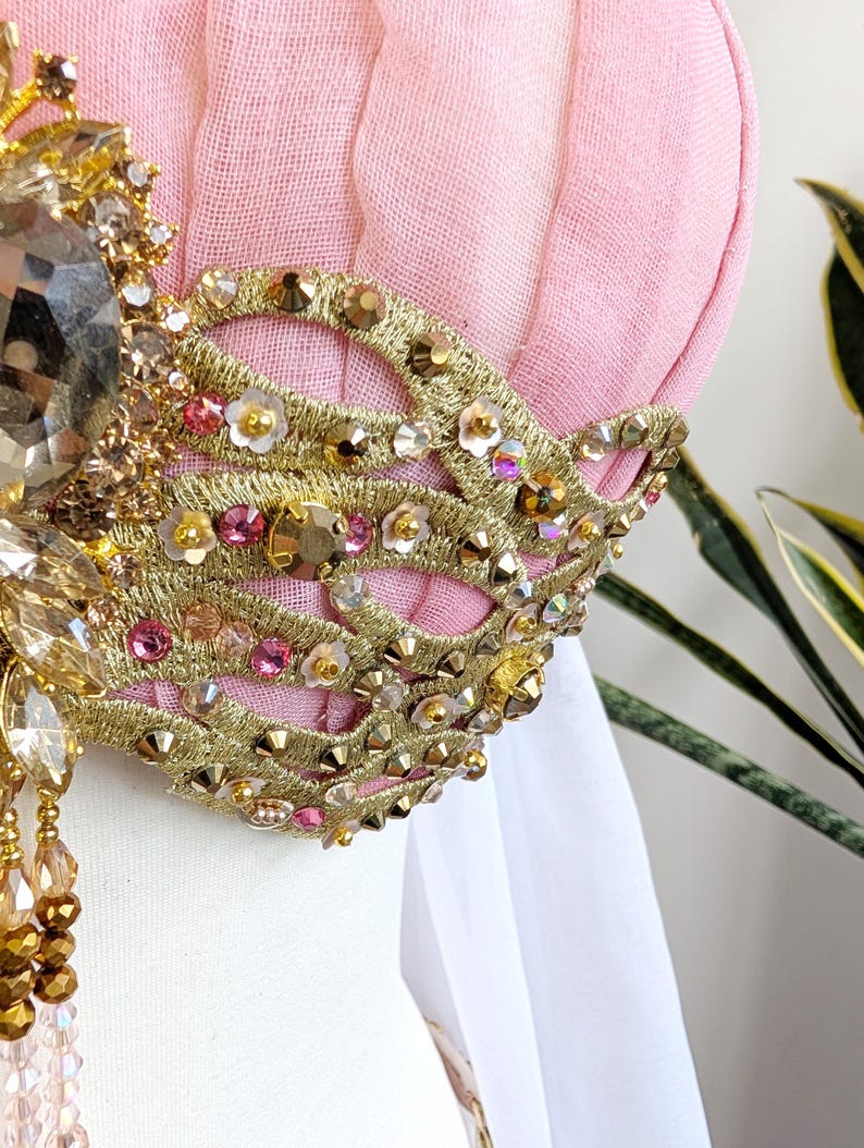 May include: A pink fabric bodice featuring intricate gold embroidery in a swirling pattern. The design is embellished with numerous small gold studs, iridescent sequins, tiny white flower appliqu&eacute;s, and pink crystals. A large faceted amber-colored crystal sits at the center, with dangling beaded strands hanging below.