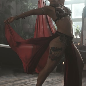 May include: A dark red, sheer, flowing skirt with a high slit, paired with a black, heavily embellished, coin-adorned bra top. The outfit includes metallic arm cuffs, a matching coin belt, and a delicate ankle bracelet, creating a layered look.