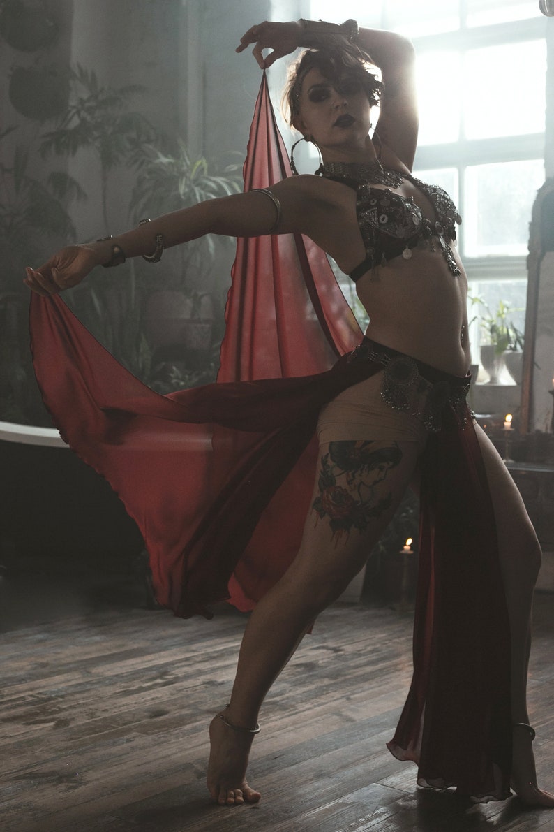 May include: A dark red, sheer, flowing skirt with a high slit, paired with a black, heavily embellished, coin-adorned bra top. The outfit includes metallic arm cuffs, a matching coin belt, and a delicate ankle bracelet, creating a layered look.