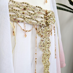 May include: A white garment featuring a wide, ornate gold belt embellished with intricate embroidery, metallic sequins, and small pink and amber beads. A central decorative panel hangs from the belt, adorned with dangling beaded fringe strands.